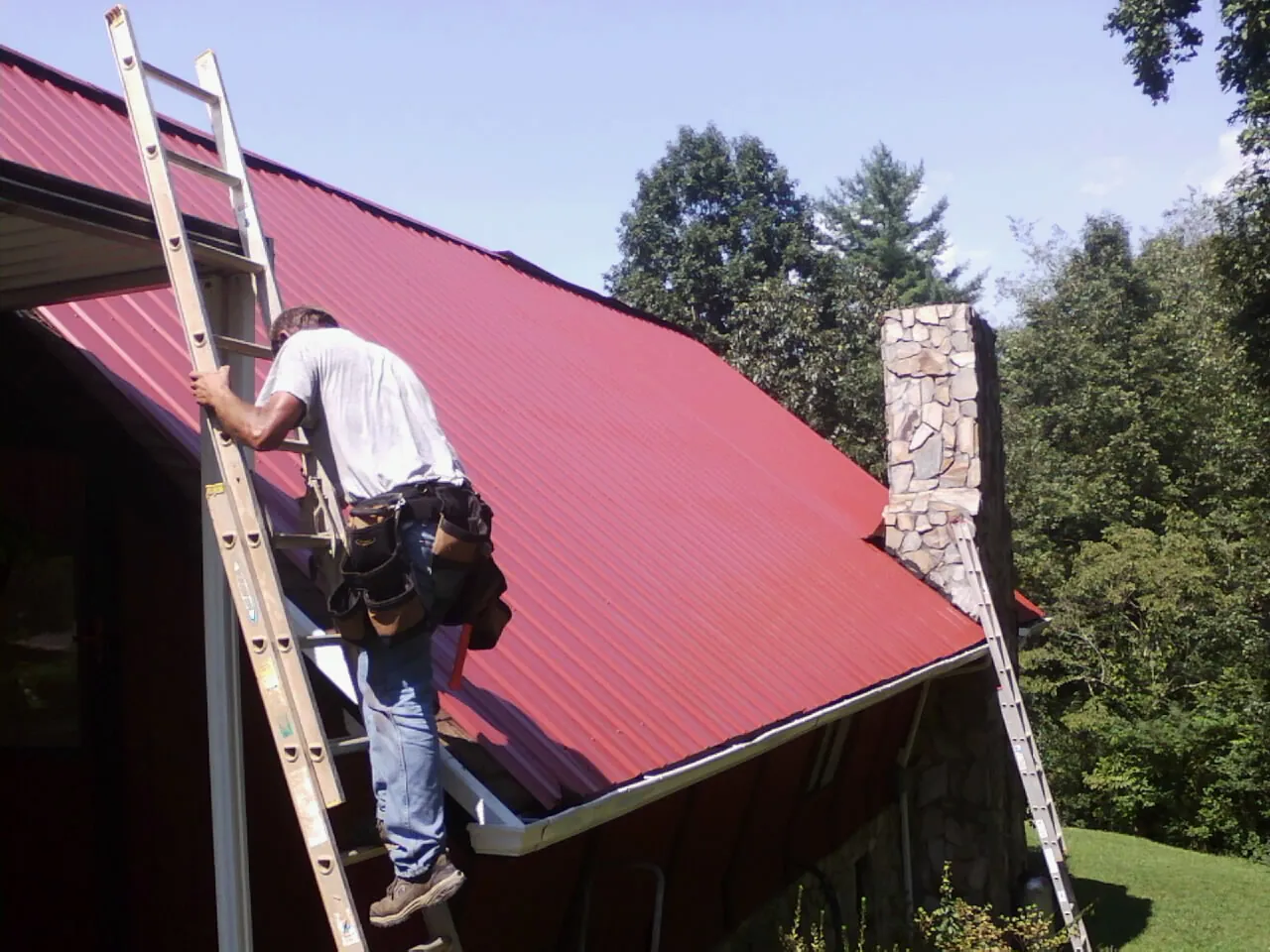 Roofer on a ladder beside a red metal roof and stone chimney — Soffit & Fascia Repair by Roofing Brothers in Timber